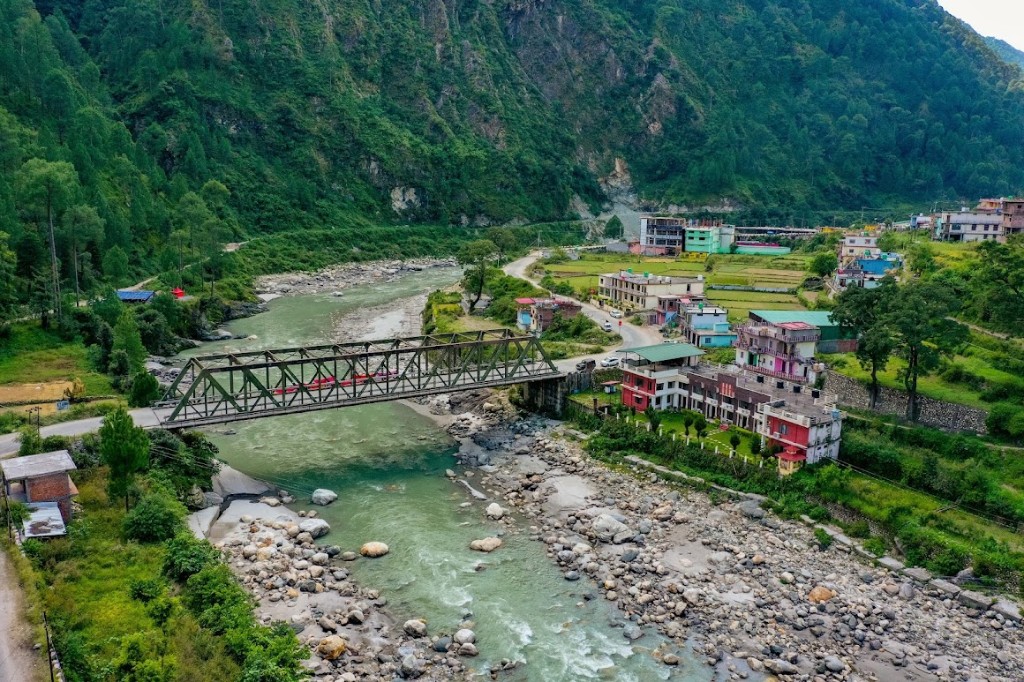 Aerial view of Shiv Parivar Resort by the river in Uttarkashi — metal bridge, turquoise water, colorful buildings, and forested Himalayan slopes